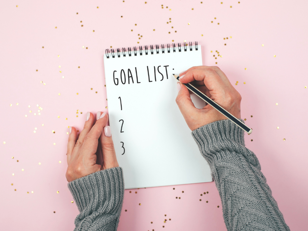 A hand writing a goal list on a pink background.